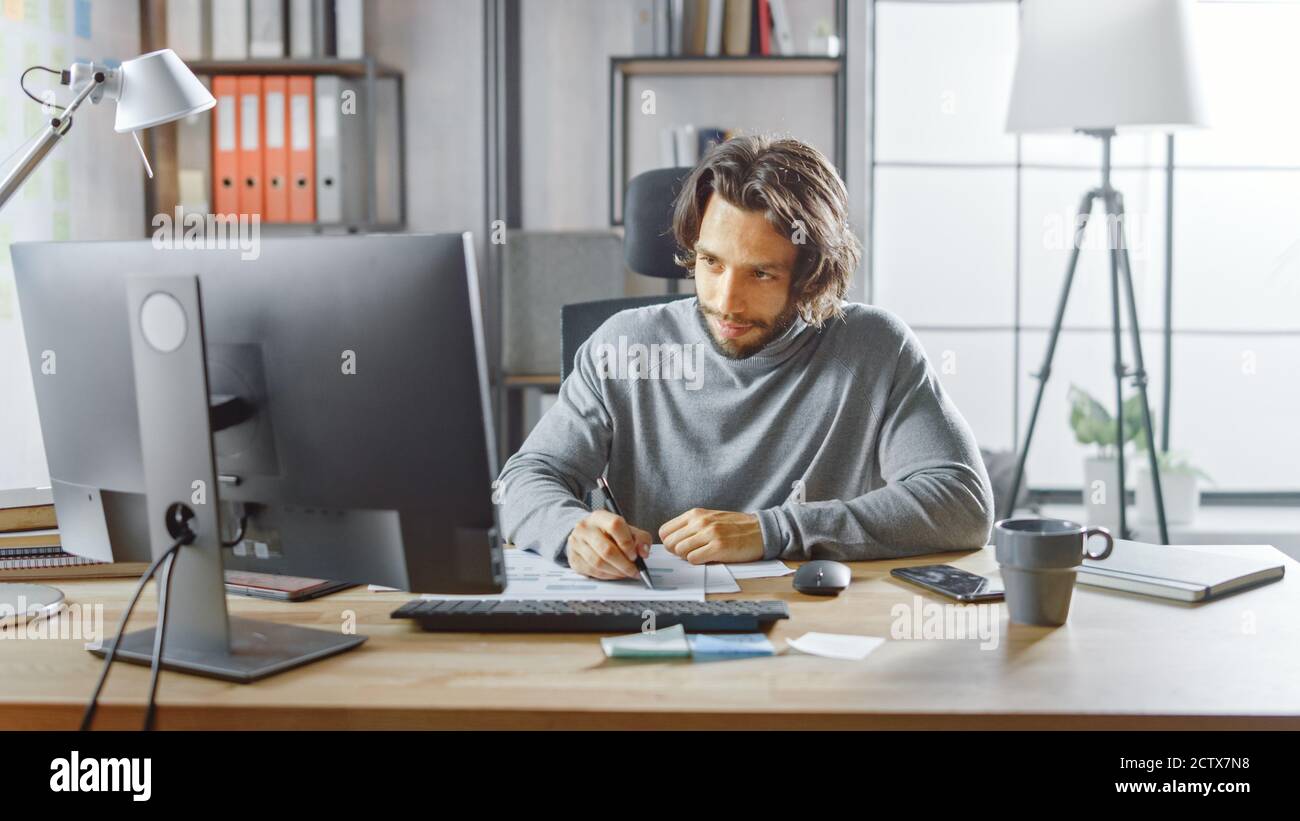 Gutaussehende langhaarige Unternehmer sitzen an seinem Schreibtisch im Büro arbeitet auf Desktop-Computer, Arbeiten mit Dokumenten, Korrigieren von Diagrammen, Zeichnen Stockfoto