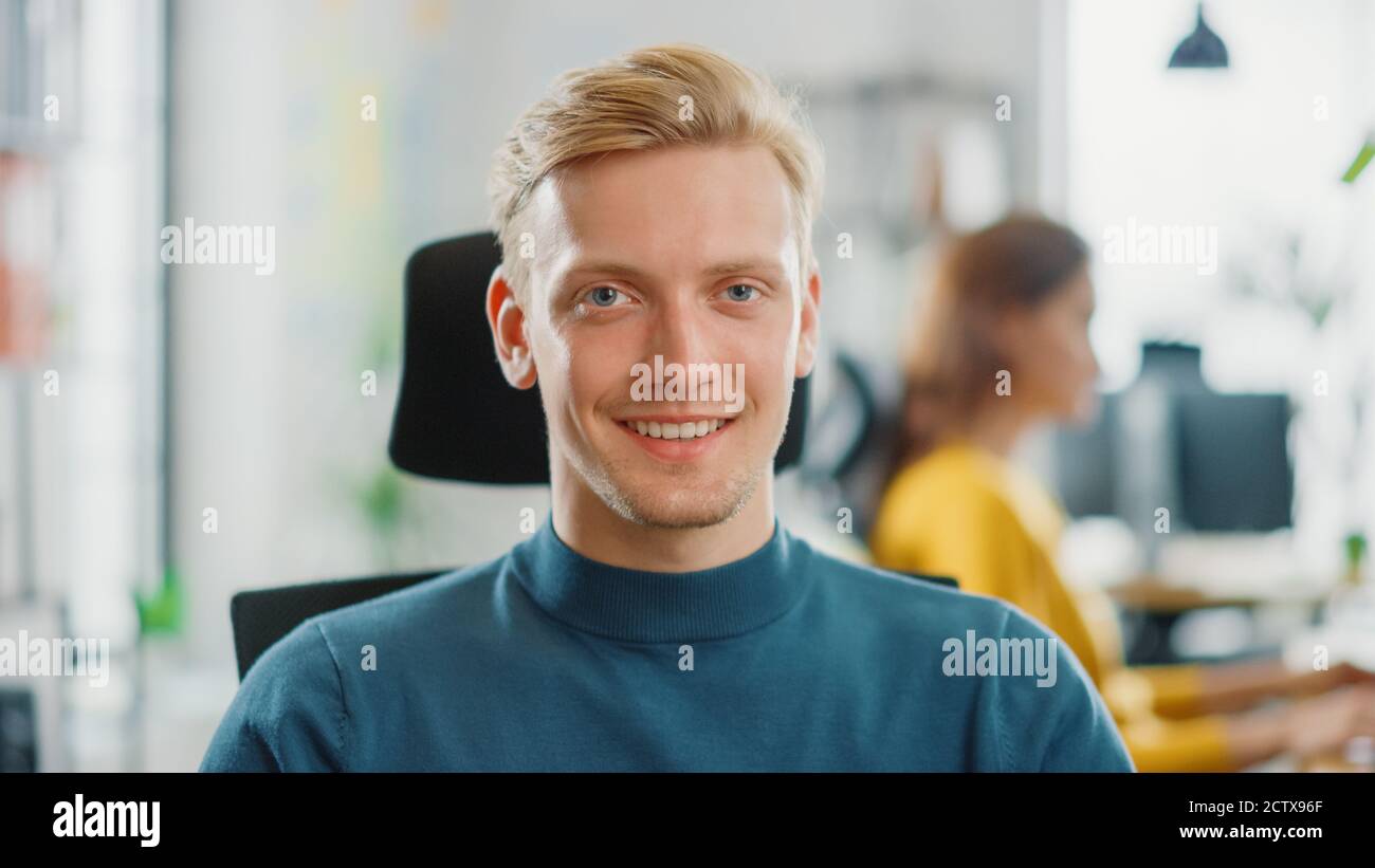Porträt eines gutaussehenden professionellen Kaukasischen Mann, der an seinem Schreibtisch arbeitet und lächelnd auf die Kamera schaut. Erfolgreicher Mann, der in Bright Diverse Office arbeitet. Stockfoto