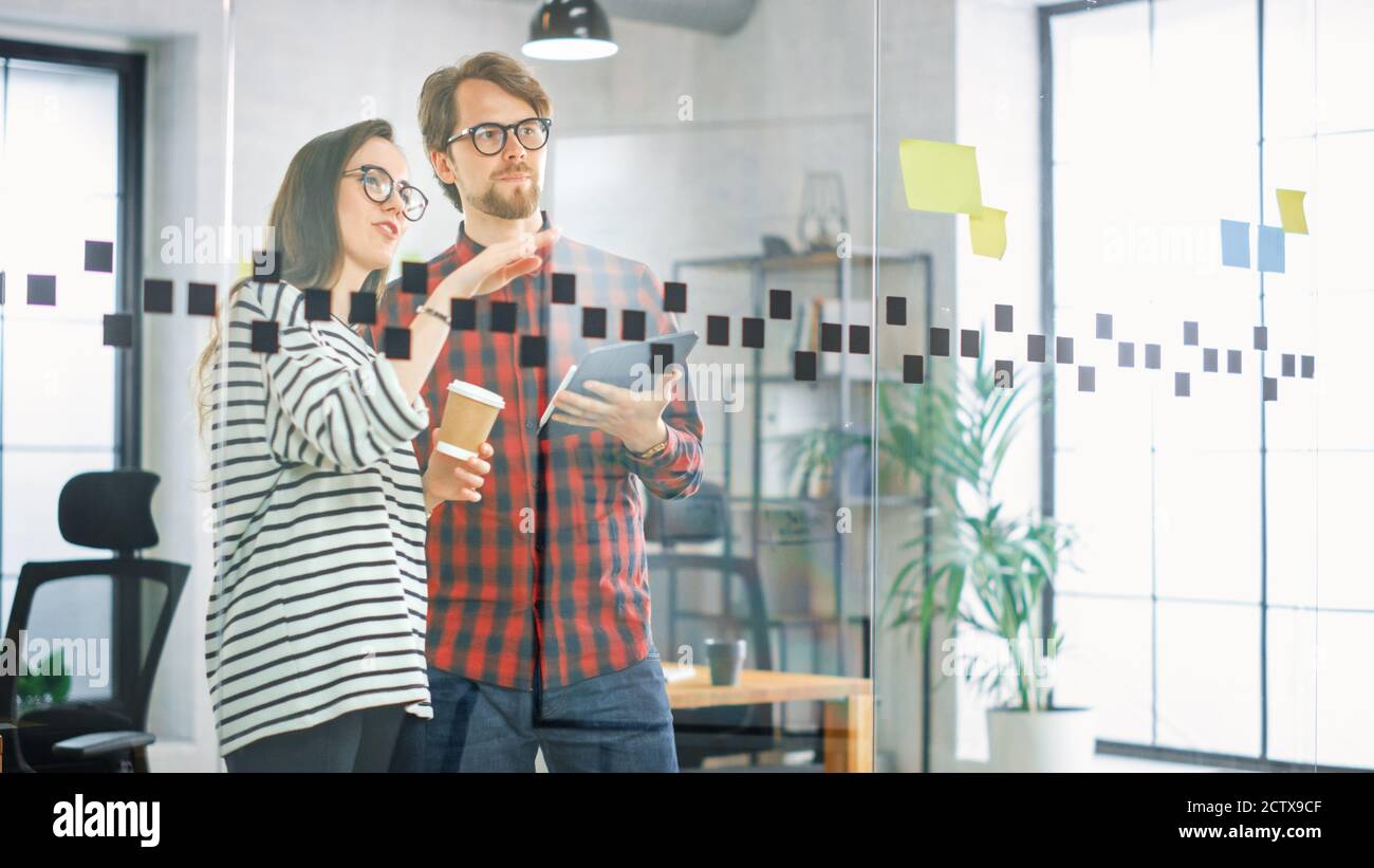 Junger Mann mit Tablet und Frau mit Takeaway Kaffeeständer vor einer Glaswand mit Notizen und diskutieren Geschäftsmöglichkeiten. Sie arbeiten in Creative Stockfoto