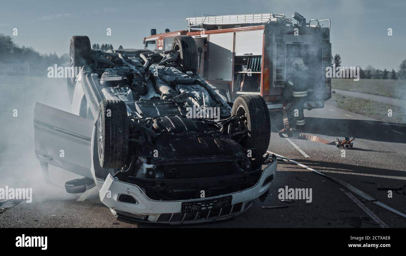 Verkehrsunfall Rollover Rauchen und Brennen Fahrzeug auf seinem Dach in der Mitte der Straße nach Kollision. Im Hintergrundteam von Stockfoto