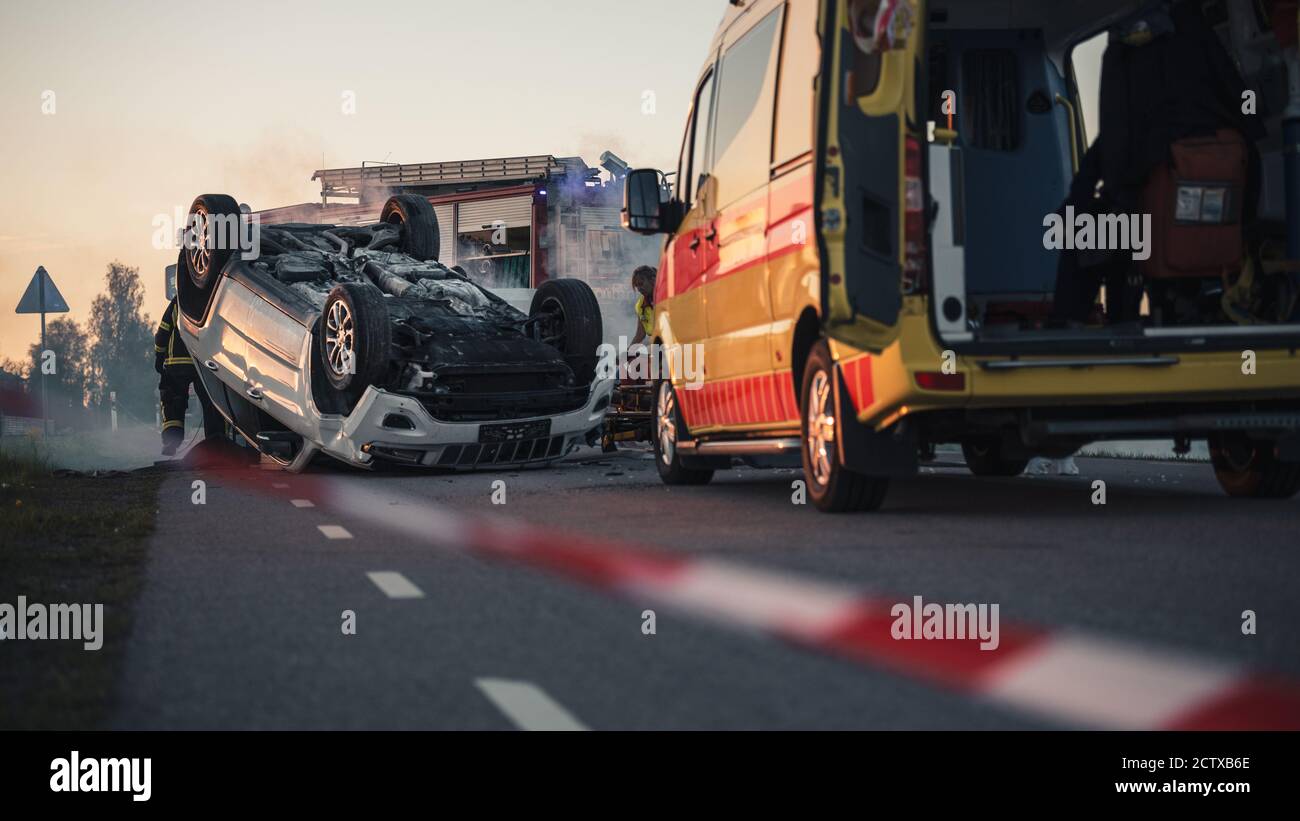 Auf dem Auto Crash Verkehr Unfallort Rollover Räuchern Fahrzeug auf seinem Dach in der Mitte der Straße nach Kollision liegen. Im Hintergrund Medics Stockfoto
