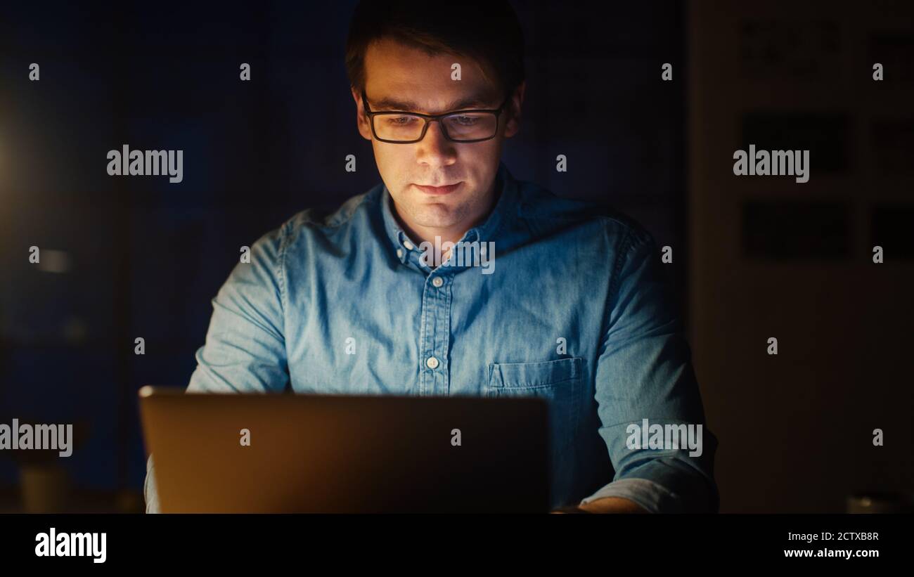 Professioneller Mann sitzt an seinem Schreibtisch im Büro Studio arbeiten auf einem Laptop am Abend. Mann, der mit Daten arbeitet, Statistiken analysiert, abschreibt Stockfoto