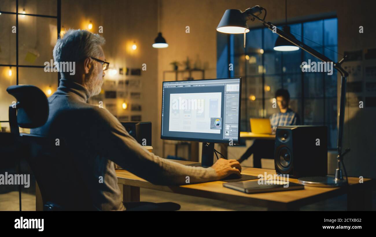 Creative Middle Aged Mobile Software Designer sitzt an seinem Schreibtisch Verwendet Desktop-Computer mit Bildschirm, auf dem der Designprozess der Smartphone-Anwendung angezeigt wird Stockfoto