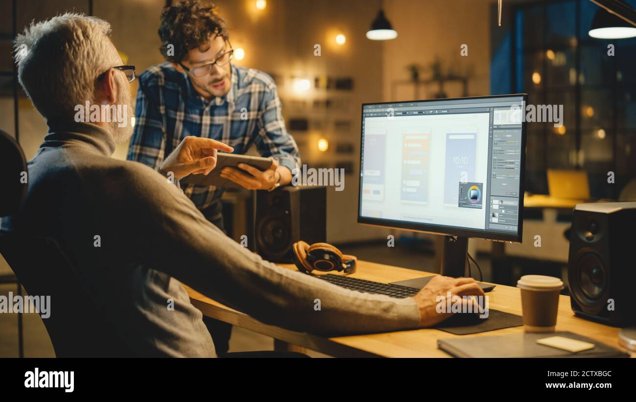 Am Abend Creative Middle Aged Mobile Software Designer nutzt Desktop-Computer mit Smartphone Application Design, hat Gespräch mit jüngeren Stockfoto