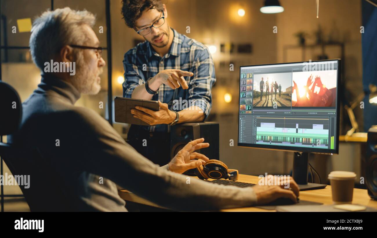 Abend in Creative Office: Professionelle Middle Aged Videographer arbeitet auf Desktop-Computer, hat Gespräch mit Younger Specialist, der Tablet verwendet Stockfoto