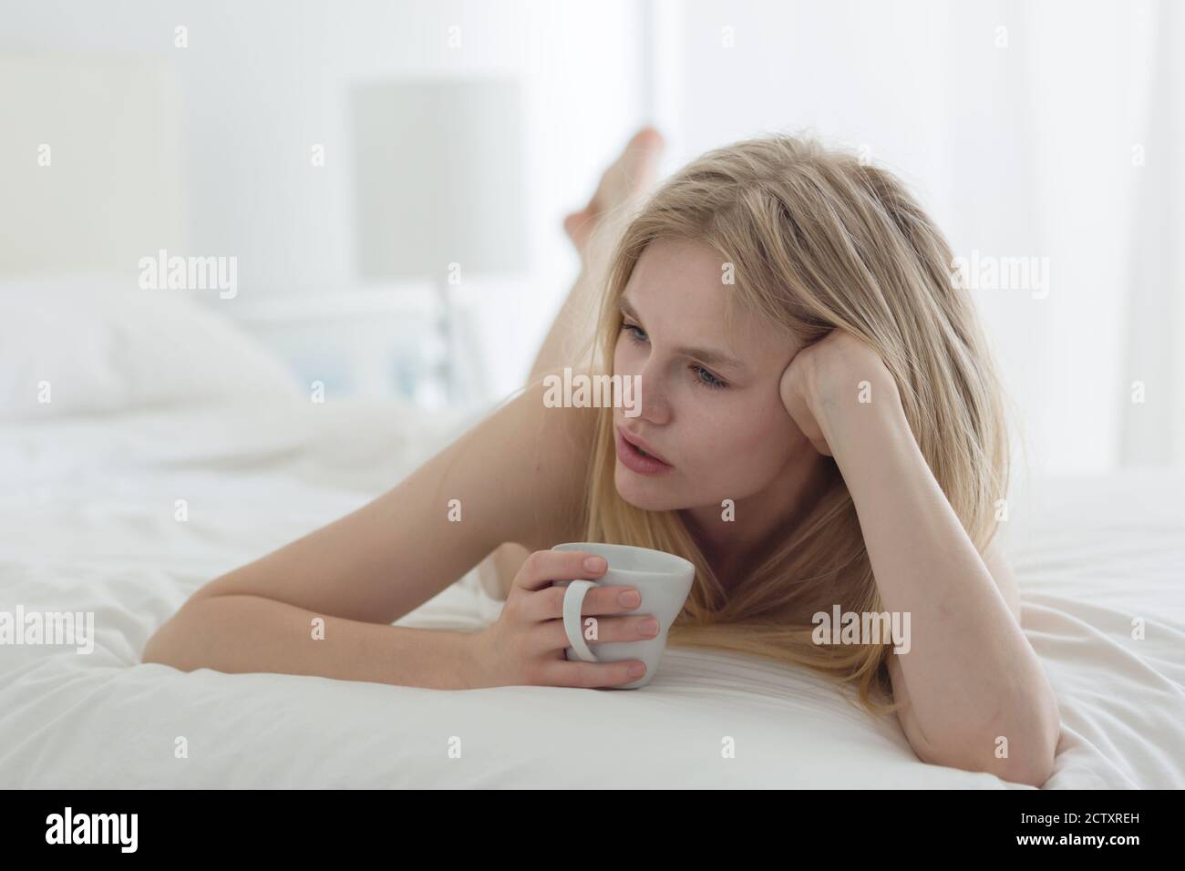 Nachdenkliche junge blonde Frau, die Kaffee trinkt, während sie im Bett liegt. Stockfoto