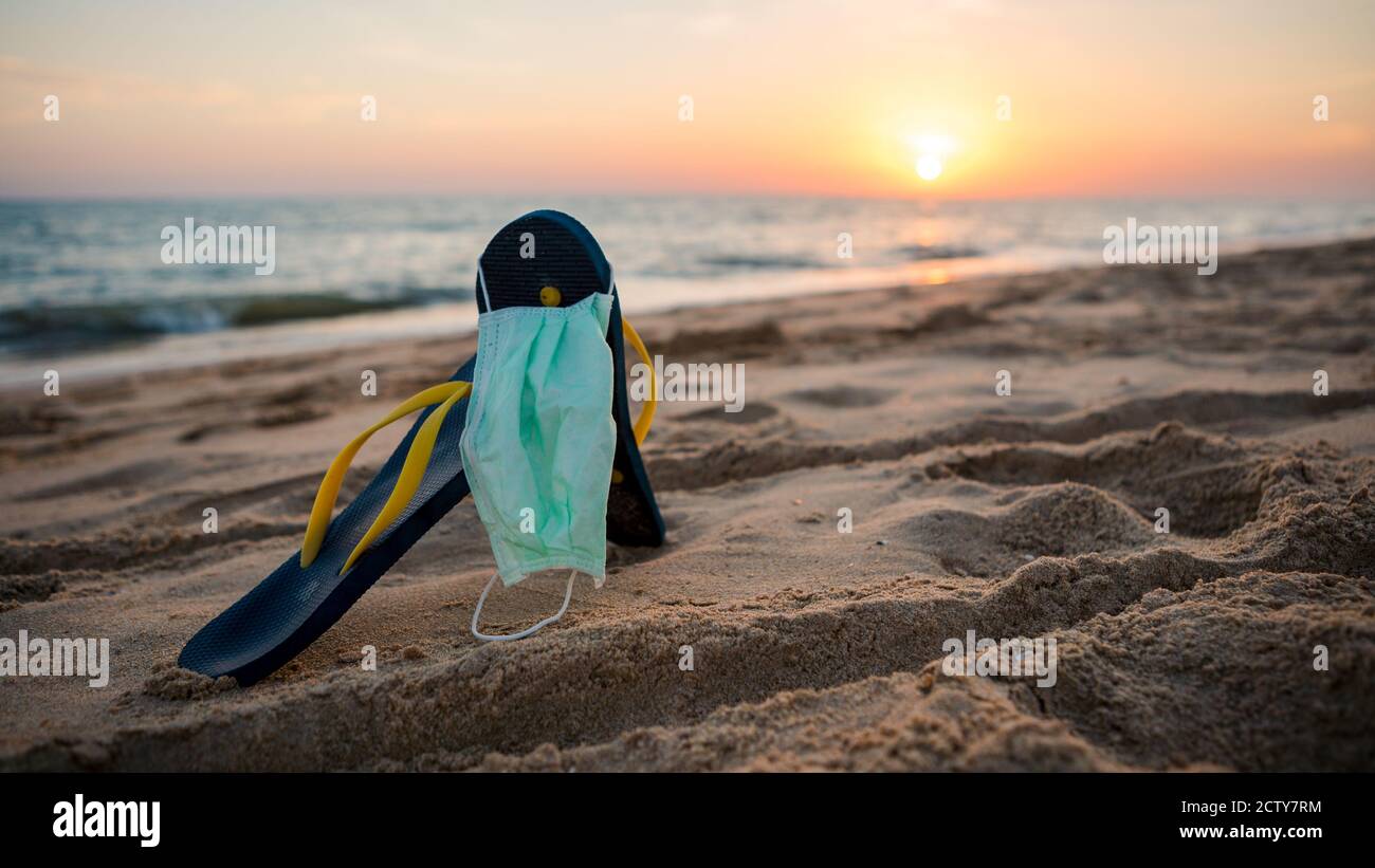 Slipper und medizinische Maske auf dem schönen Strand mit Sonnenuntergang orange Licht. Schutzmaske für COVID-19 mit Sandale an der spanischen Küste. Urlaub und unloc Stockfoto
