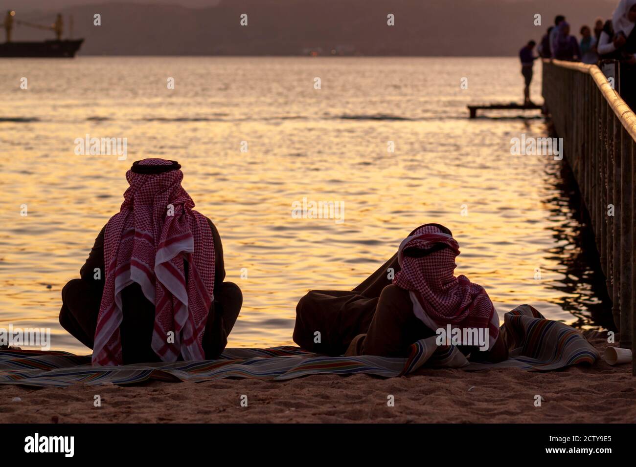 Zwei arabische Männer mit Keffiyeh, Agal und Thobe sind am Strand neben einem Pier zu sehen. Einer liegt auf dem Rücken, der andere sitzt mit gekreuzten Beinen. Sie Stockfoto