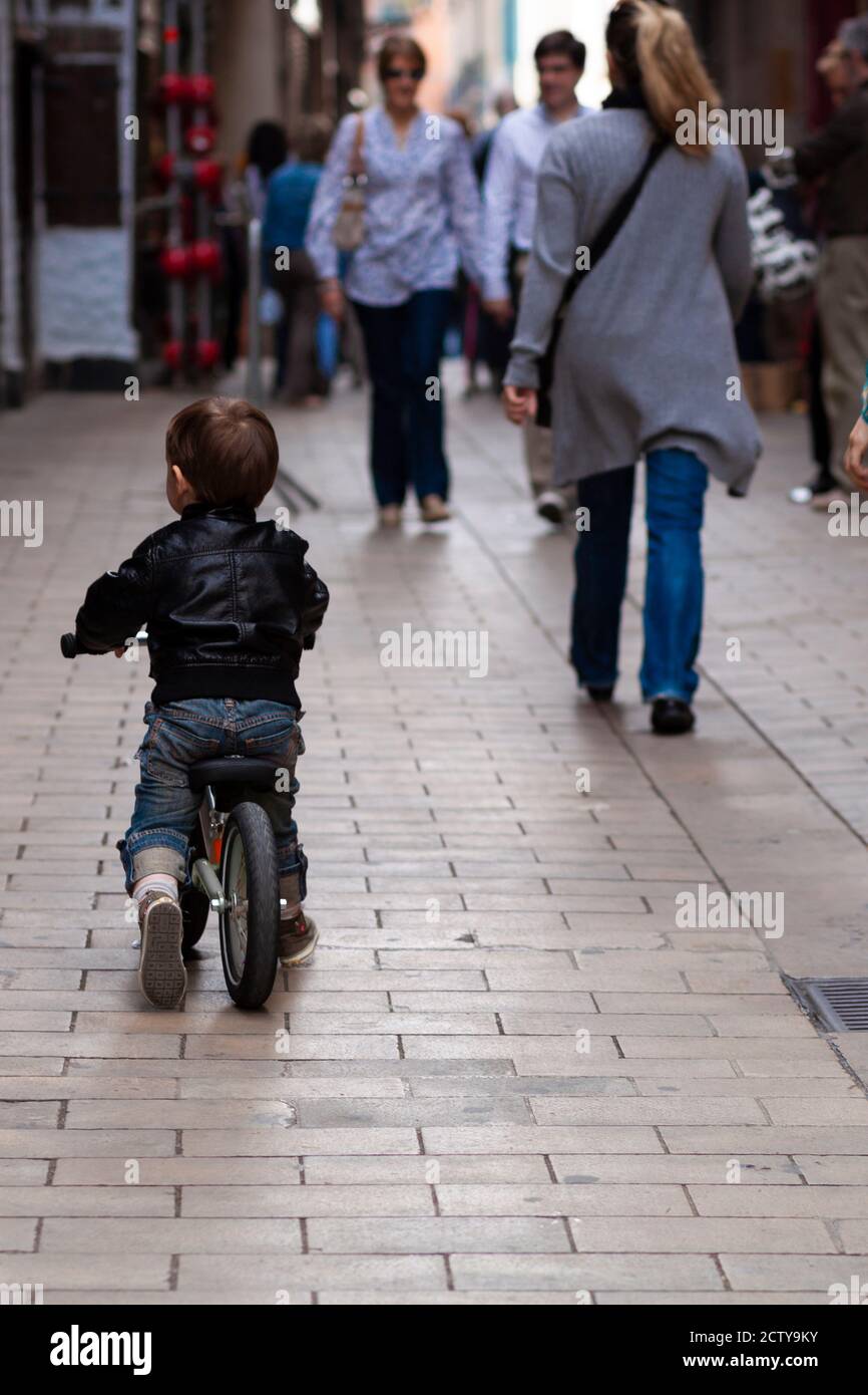 Nahaufnahme Bild zeigt einen niedlichen kleinen Jungen trägt coole stilvolle Jeans und eine Lederjacke, ein Fahrrad fahren in einer belebten Straße mit Geschäften und Menschen vorbei Stockfoto