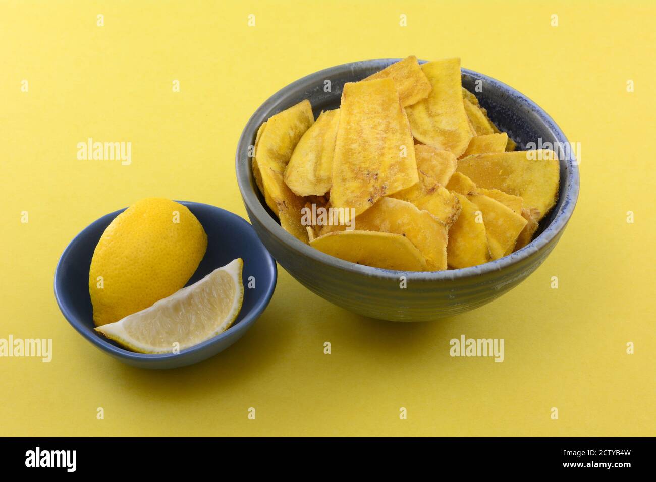 Kochbananen-Chips mit Zitronengeschmack in einer Snack-Schüssel mit Zitronenkeilen In blauer Schale auf gelbem Hintergrund Stockfoto