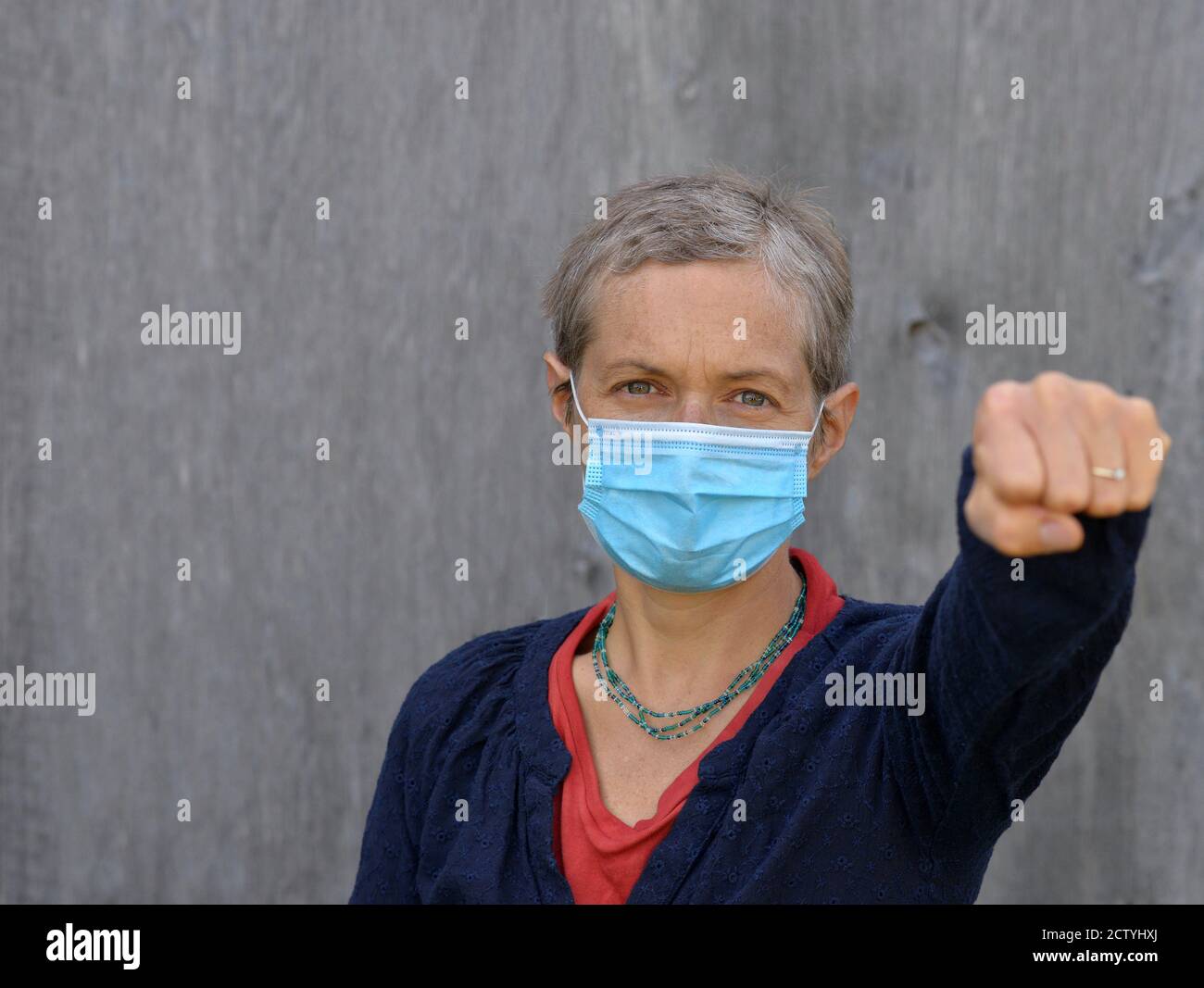 Die kaukasische Frau mittleren Alters mit kurzen Haaren trägt eine chirurgische Gesichtsmaske und zeigt während der Corona-Virus-Pandemie von 2019/20 ihre Faust in Richtung Kamera. Stockfoto