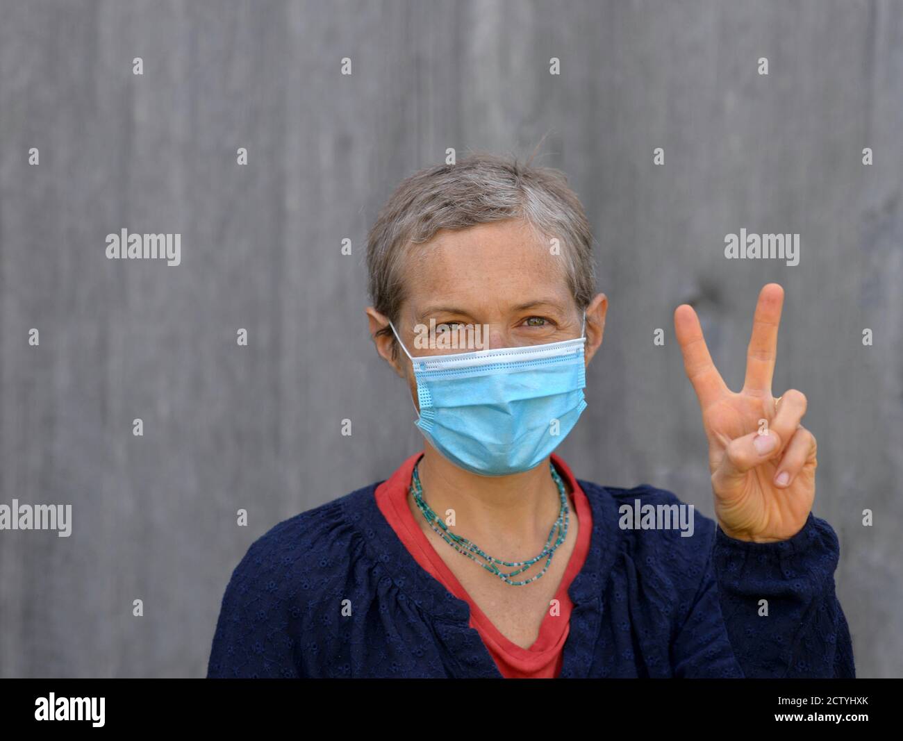 Kaukasische Frau mittleren Alters mit kurzen Haaren trägt eine blaue Einweg-OP-Gesichtsmaske und macht das V-Zeichen während der 2019/20 Corona-Virus Pandemie. Stockfoto