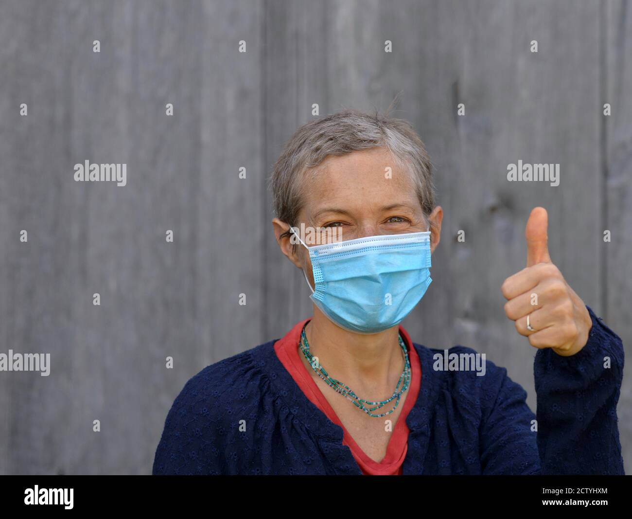 Die kaukasische Frau mittleren Alters mit kurzen Haaren trägt eine blaue chirurgische Gesichtsmaske und gibt während der Corona-Virus-Pandemie von 2019/20 das Daumen-up-Zeichen. Stockfoto