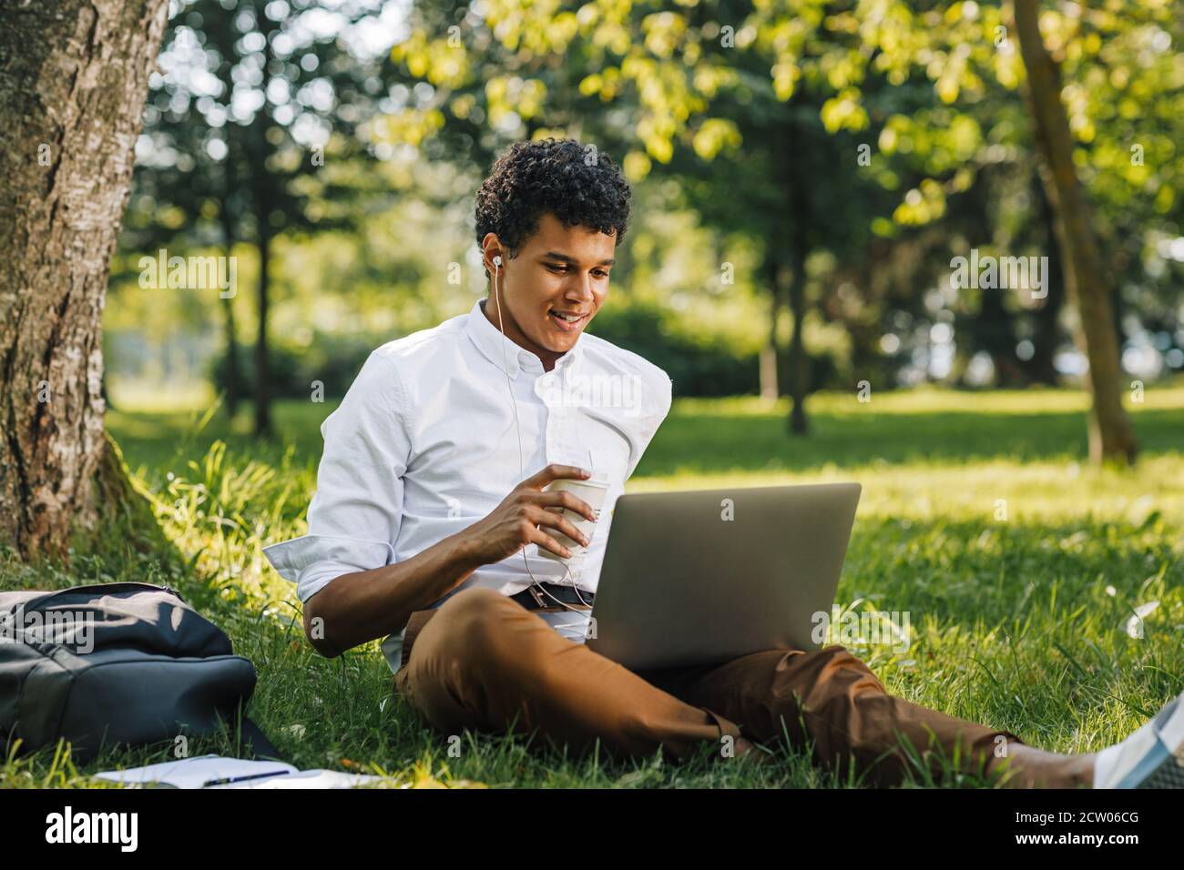 Lächelnder afroamerikanischer College-Junge mit einem Laptop im Park sitzend. Junger Mann, der auf dem Laptop schaut und Kaffee trinkt. Stockfoto
