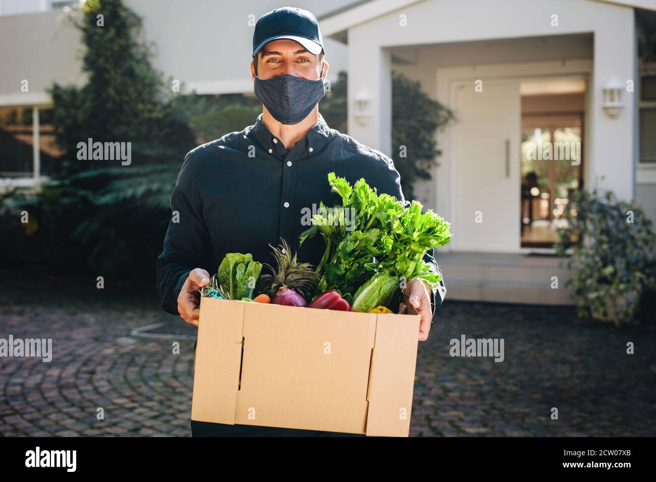 Angestellter trägt eine Gesichtsmaske und hält einen Lebensmittelkarton draußen. Mann, der eine Schachtel Obst und Gemüse an die Adresse nach Hause liefert. Stockfoto