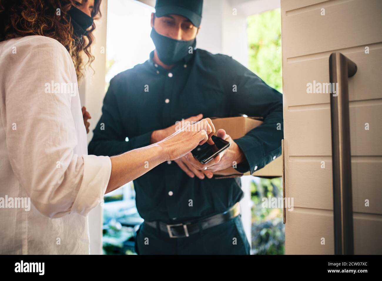 Delivery Mann mit Gesichtsmaske hält Smartphone, während weibliche Kunden Unterzeichnung auf Touchscreen Empfang von Haus Lieferung Paket. Stockfoto