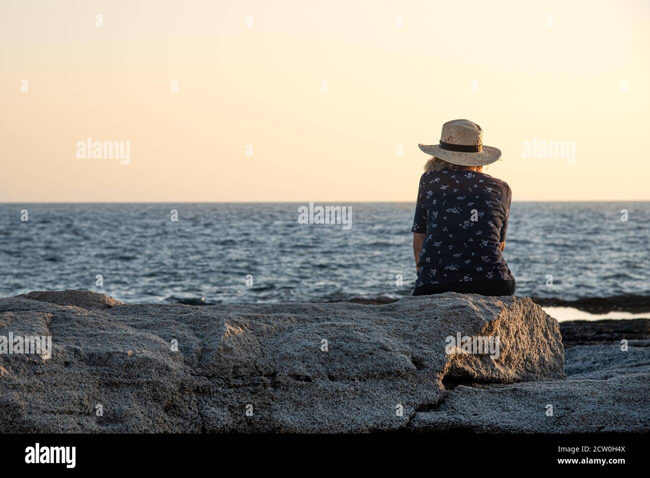 Unerkannte Frau, die auf einem Felsen sitzt und das Meer bei Sonnenuntergang genießt. Stockfoto
