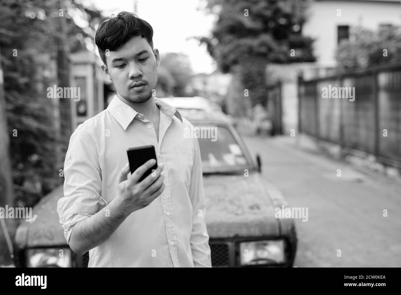 Junge asiatische Geschäftsmann mit Mobiltelefon gegen rostigen Auto Stockfoto