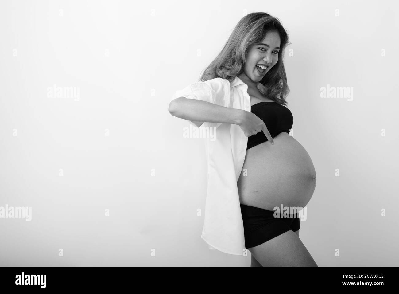 Studio Aufnahme von jungen glücklich asiatische schwangere Frau lächeln während Zeigt auf ihren Bauch und trägt ein offenes weißes Hemd Stockfoto