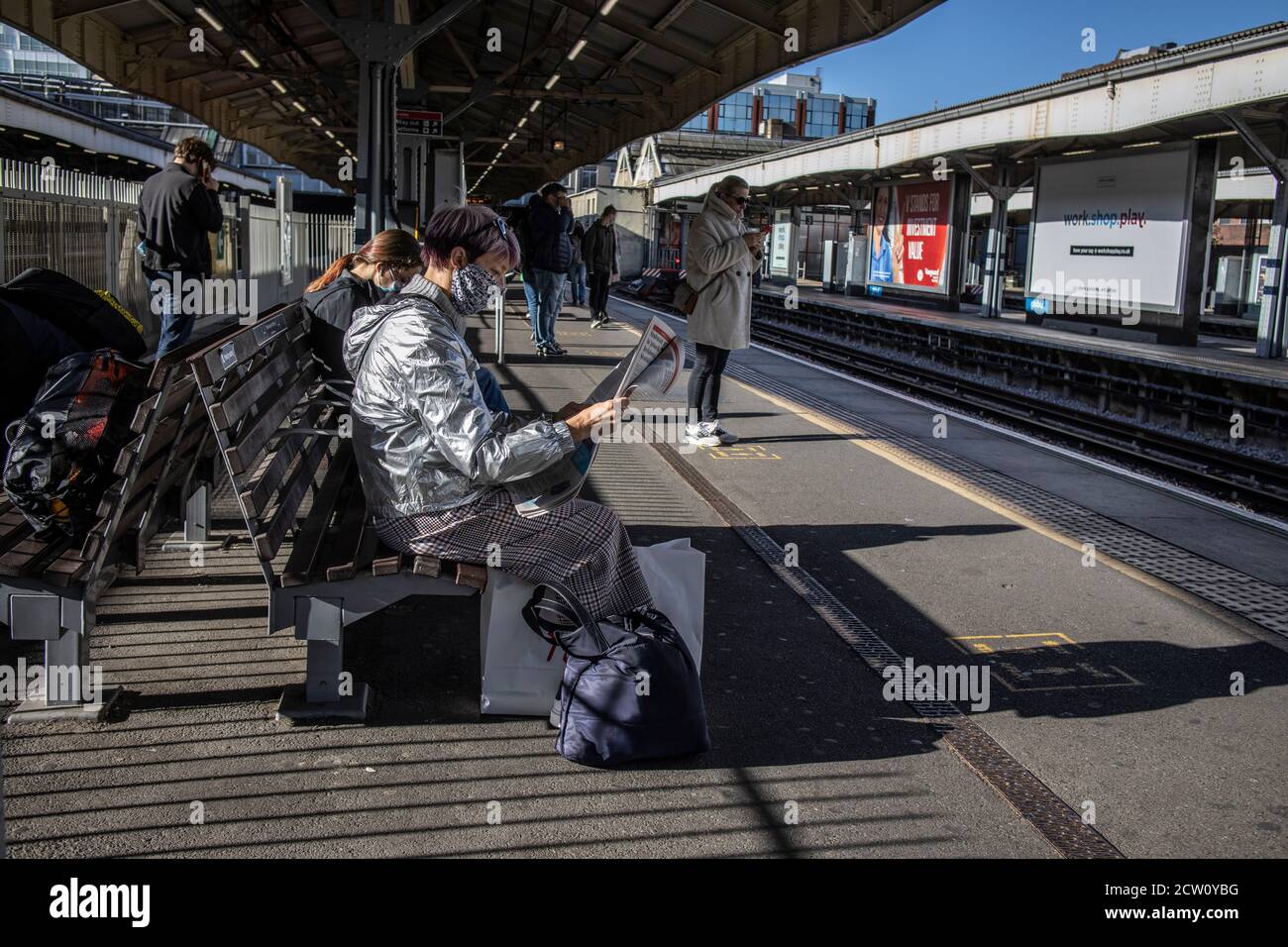 Frau mit silberfarbenem wasserdichtem Mantel liest eine kostenlose Zeitung auf der Plattform der Wimbledon Station, Südwestengland, Großbritannien Stockfoto
