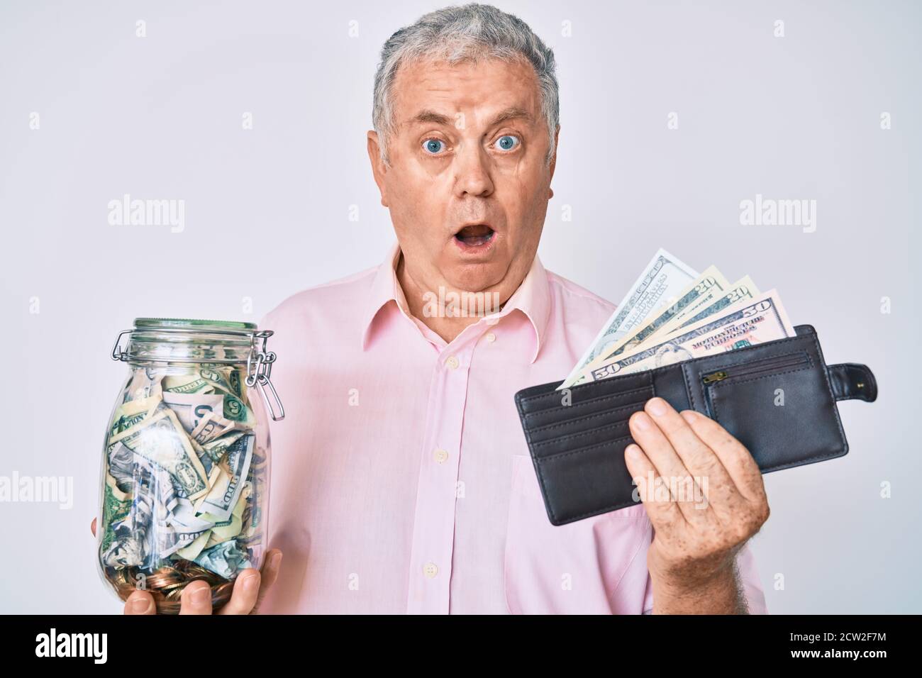 Älterer grauhaariger Mann hält Geldbörse mit Dollar und Glas mit Ersparnissen im Schockgesicht, sieht skeptisch und sarkastisch aus, überrascht mit offenem Mund Stockfoto