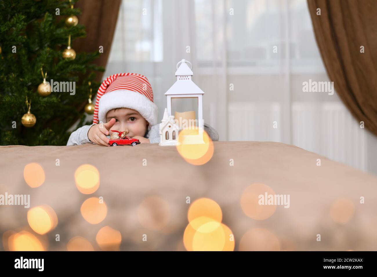 Ein Junge in einer gestreiften Mütze gekleidet spielt mit einem roten Auto, schiebt, in der Nähe des Weihnachtsbaumes in der Nähe der Laterne mit einem Haus. Stockfoto