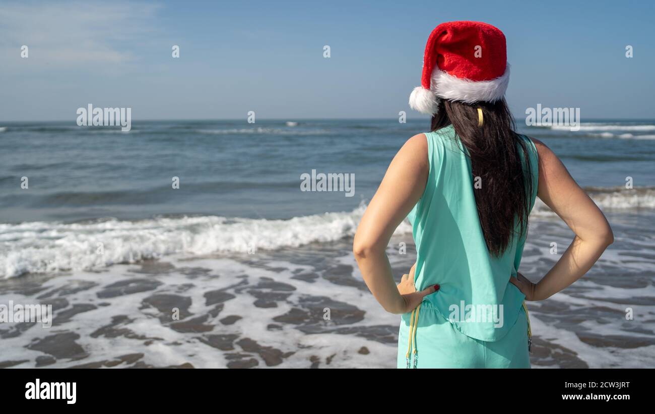 Rückansicht einer jungen Frau im Weihnachtsmann-Hut, die an einem sonnigen Tag am Meer steht und in die Ferne blickt. Erwachsene Brünette im Weihnachtshut feiern Stockfoto