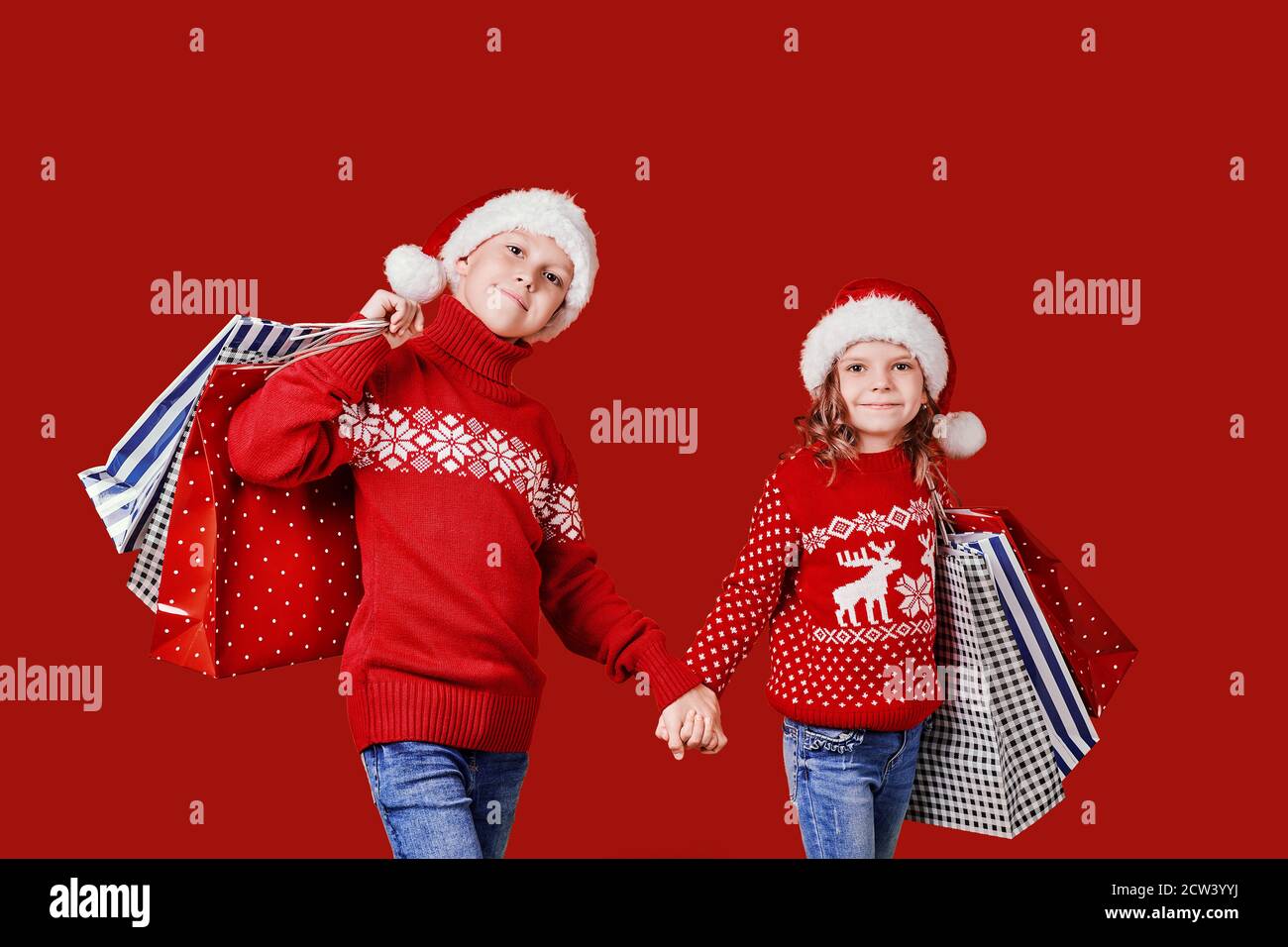 Niedliche Kinder in roten Weihnachtsmützen, Pullover mit Einkaufstaschen auf rotem Hintergrund. Stockfoto