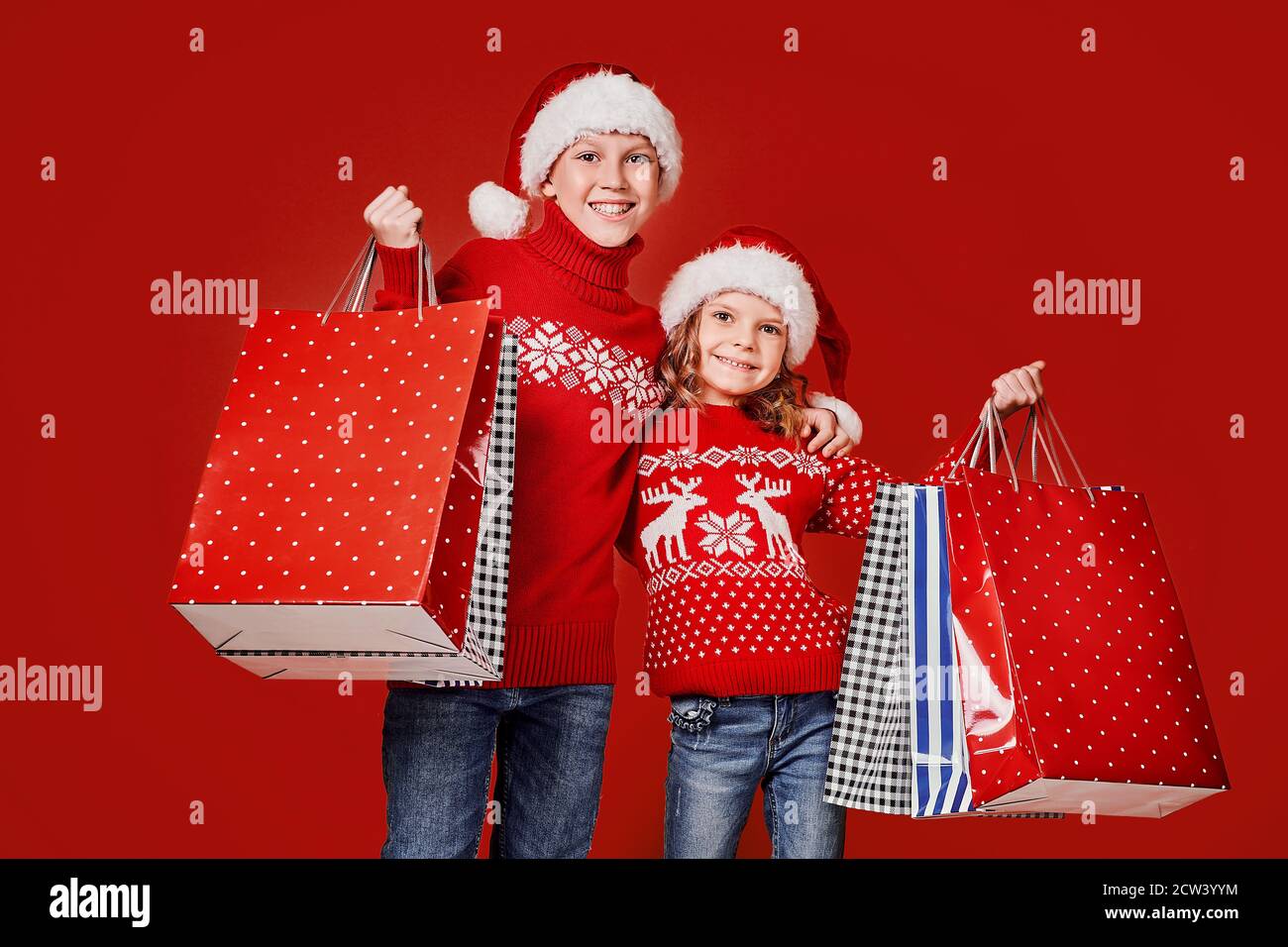 Niedliche Kinder in roten Weihnachtsmützen, Pullover mit Einkaufstaschen auf rotem Hintergrund. Stockfoto