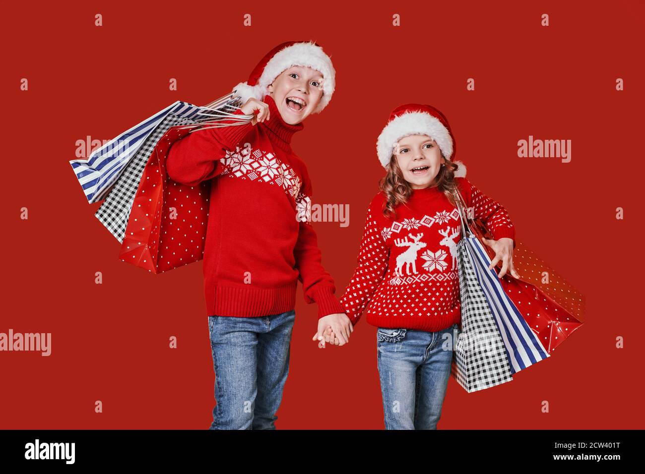 Niedliche Kinder in roten Weihnachtsmützen, Pullover mit Einkaufstaschen auf rotem Hintergrund. Stockfoto