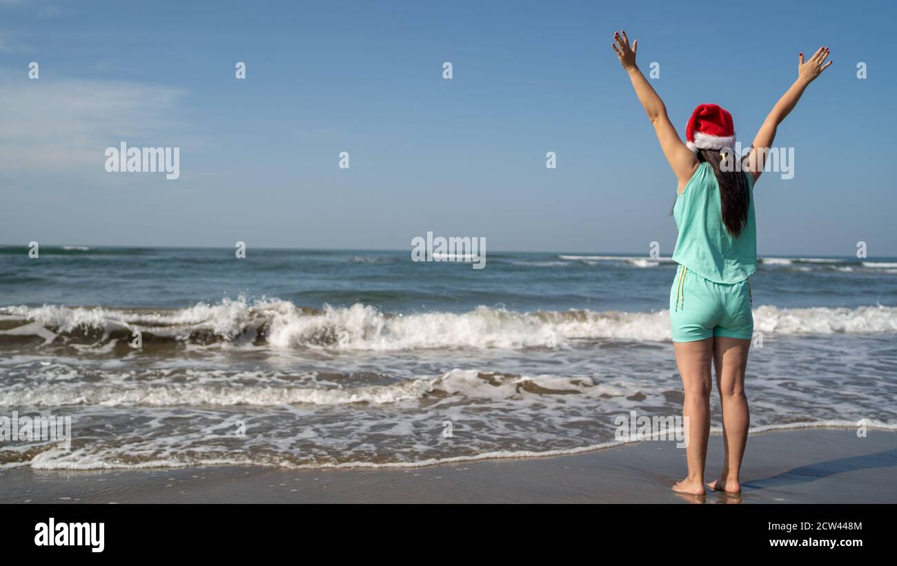 Rückansicht einer jungen Frau im Weihnachtsmann-Hut, die an einem sonnigen Tag am Meer steht und in die Ferne blickt. Erwachsene Brünette im Weihnachtshut feiern Stockfoto