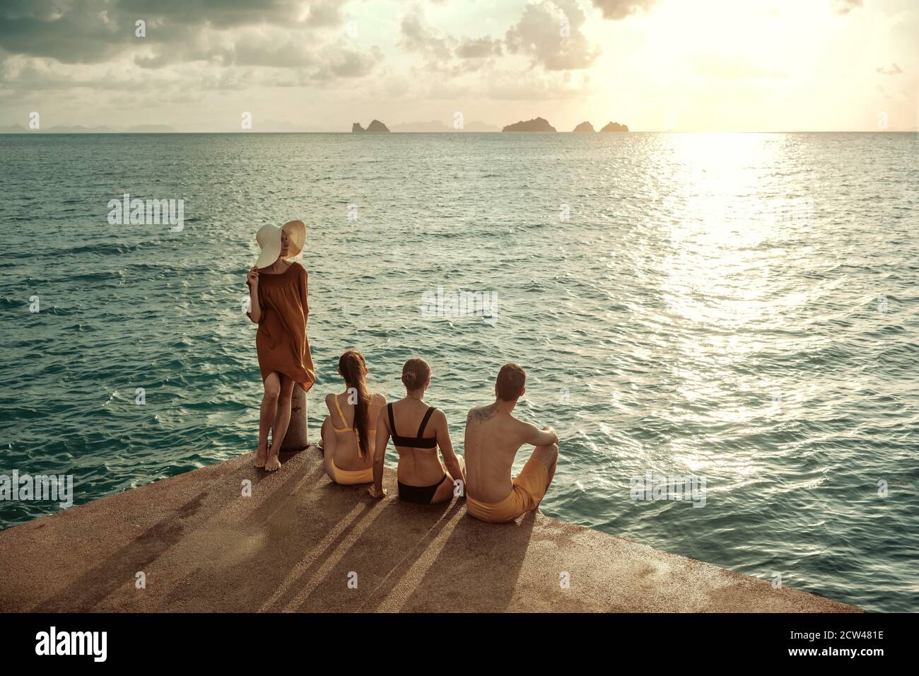 Vier junge Leute sitzen am Pier und blicken auf das Meer Und Sonnenuntergang Stockfoto