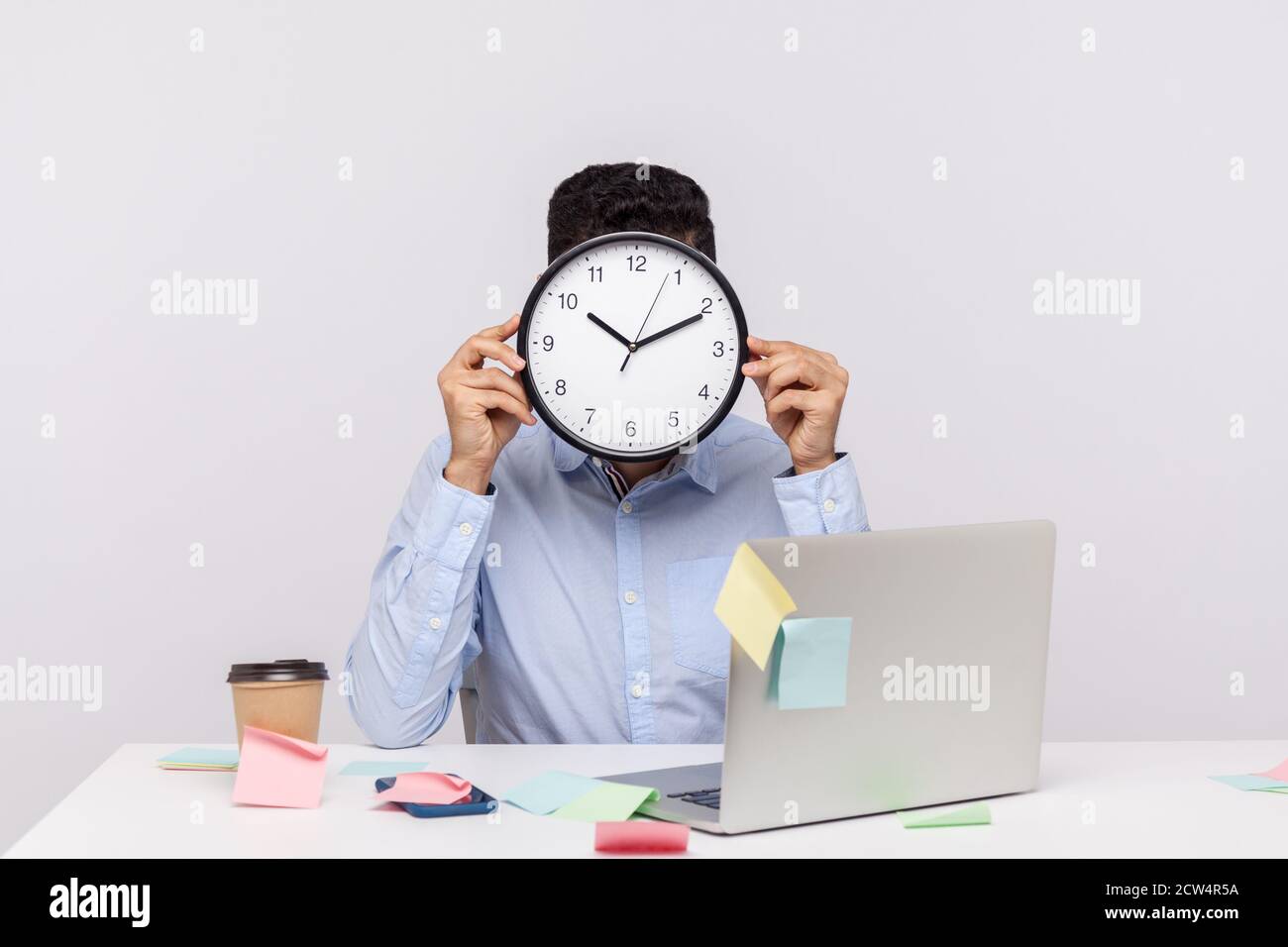Überstunden. Anonymer Mann Mitarbeiter sitzt im Büro Arbeitsplatz, versteckt Gesicht hinter der Uhr, klebrige Notizen rund um erinnern an Termin, stressig Stockfoto