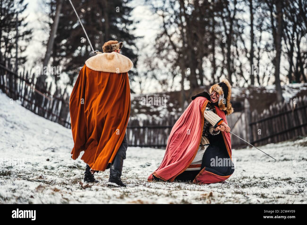 Schlacht von zwei Rittern in farbigen Gewändern und Hüten in der Nähe von Wald und hölzerne Festung. Ritter kämpfen im Winter, im Schnee. Stockfoto