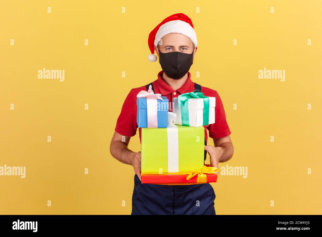 Deliveryman in schwarzer Schutzmaske und weihnachtsmann Hut in Uniform stehend und hält Pakete in den Händen, Lieferservice während Coronavirus. Innen Stockfoto
