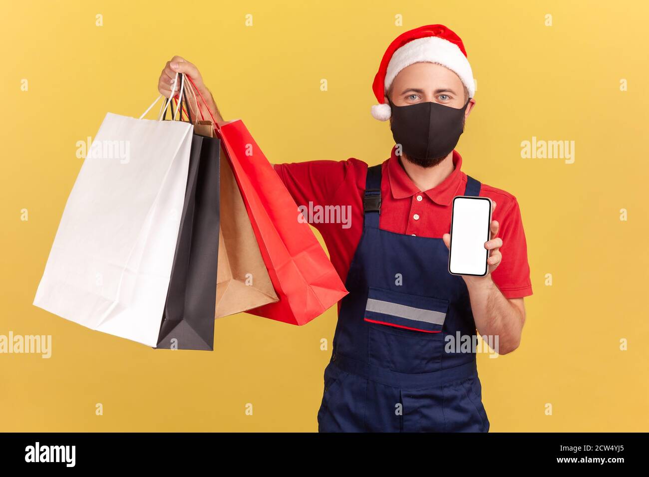 Männliche Kurier in schützende medizinische Maske und santa claus roten Hut hält Einkaufstaschen und Smartphone, einfache Bestellung und Lieferung App, Quarantäne-Dienste Stockfoto