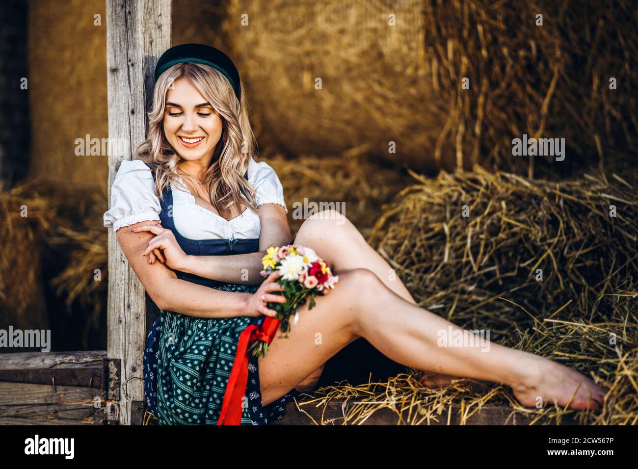 Niedliche Blondine im Dirndl, traditionelles Festkleid mit Blumenstrauß auf dem Holzzaun auf dem Bauernhof mit Heuhaufen Stockfoto