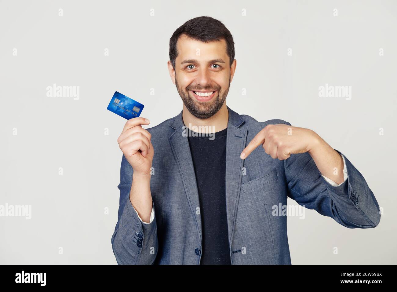 Junge Geschäftsmann Mann mit Bart in Jacke hält Kreditkarte auf isoliertem weißen Hintergrund mit überrascht Gesicht zeigt mit Finger auf sich. Porträt eines Mannes auf grauem Hintergrund. Stockfoto