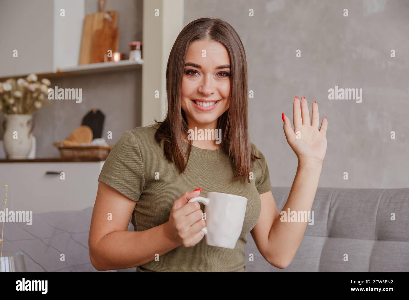 Porträt einer angenehmen jungen Frau mit Tasse Blick auf die Kamera, winken Hallo. Hübsche Millennial Dame, die per Videoanruf mit den Eltern kommuniziert Stockfoto