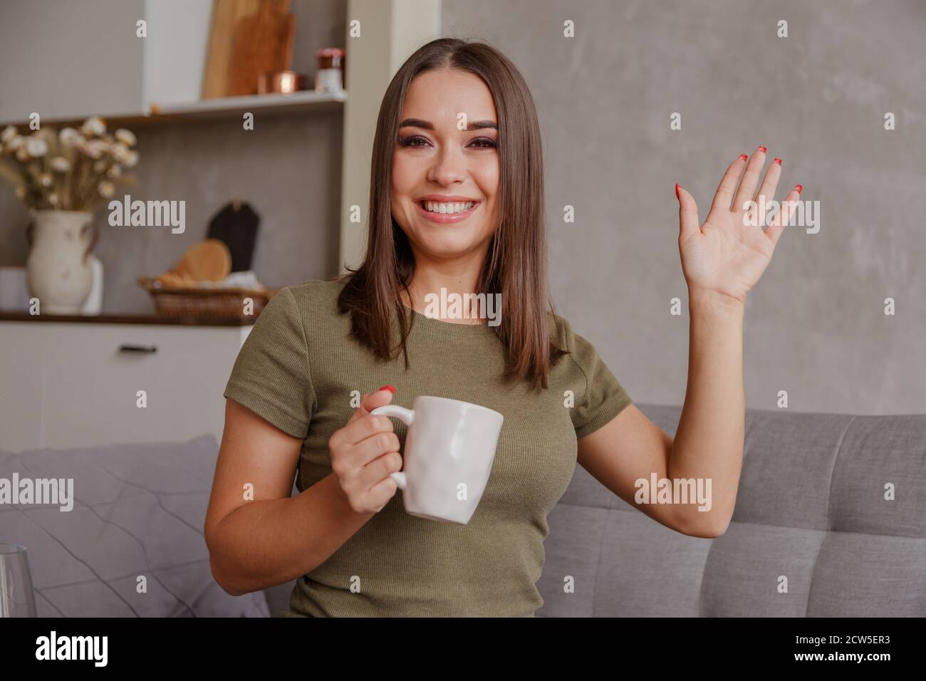 Porträt einer angenehmen jungen Frau mit Tasse Blick auf die Kamera, winken Hallo. Hübsche Millennial Dame, die per Videoanruf mit den Eltern kommuniziert Stockfoto