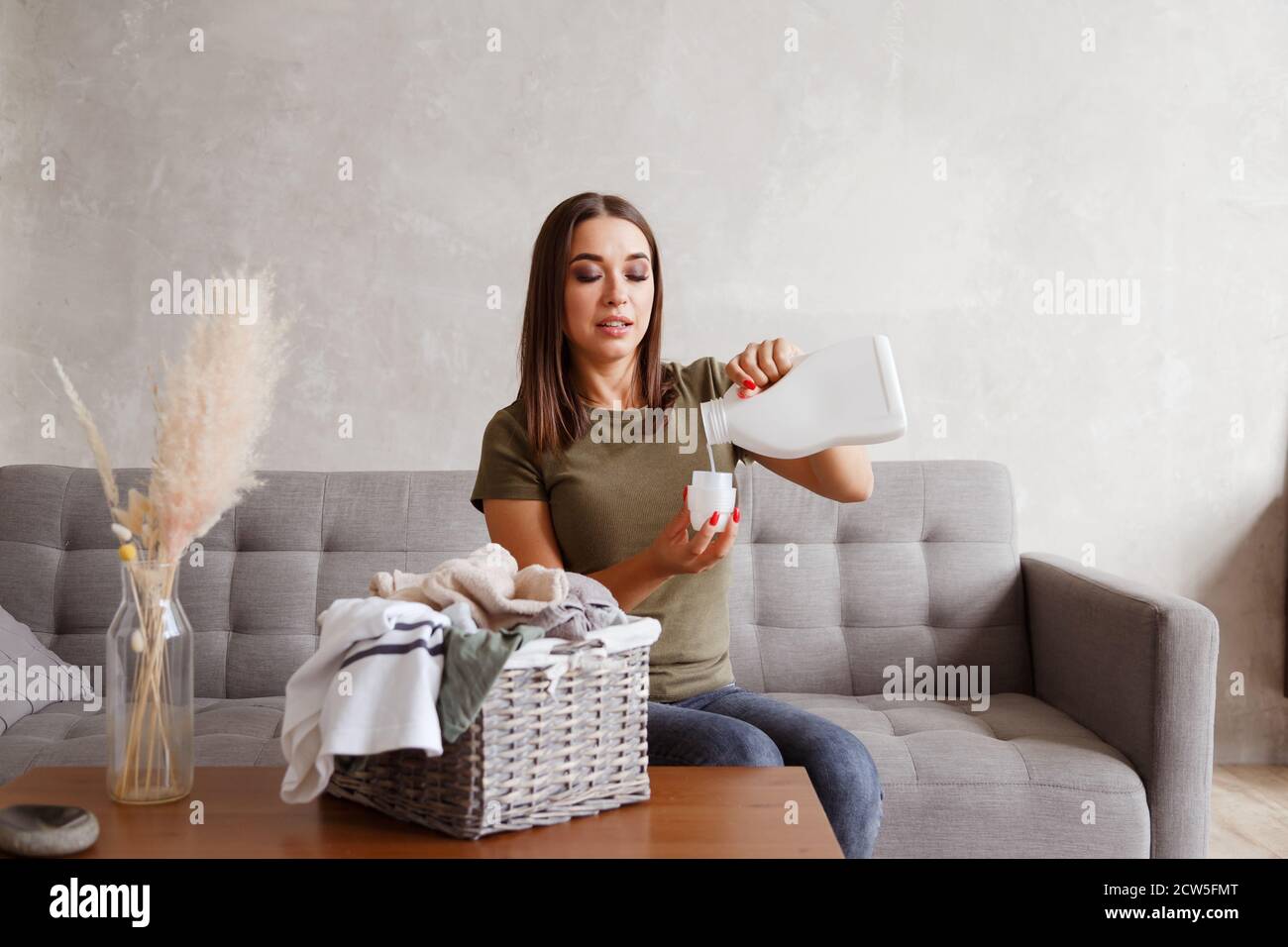 Frau gießt Waschmittel in den Flaschendeckel. Sie sitzt am Tisch mit Kleidung, die im Korb gesammelt ist Stockfoto