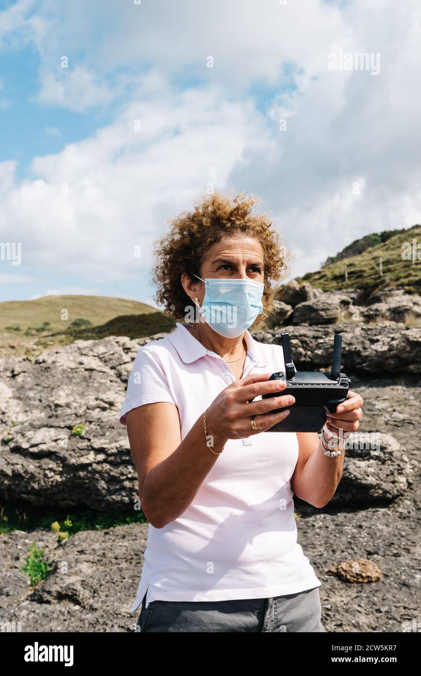 Frau mittleren Alters trägt Gesichtsmaske Navigation einer fliegenden Drohne mit Fernbedienung. Frau in der Natur Drohne fliegen und fotografieren. Stockfoto