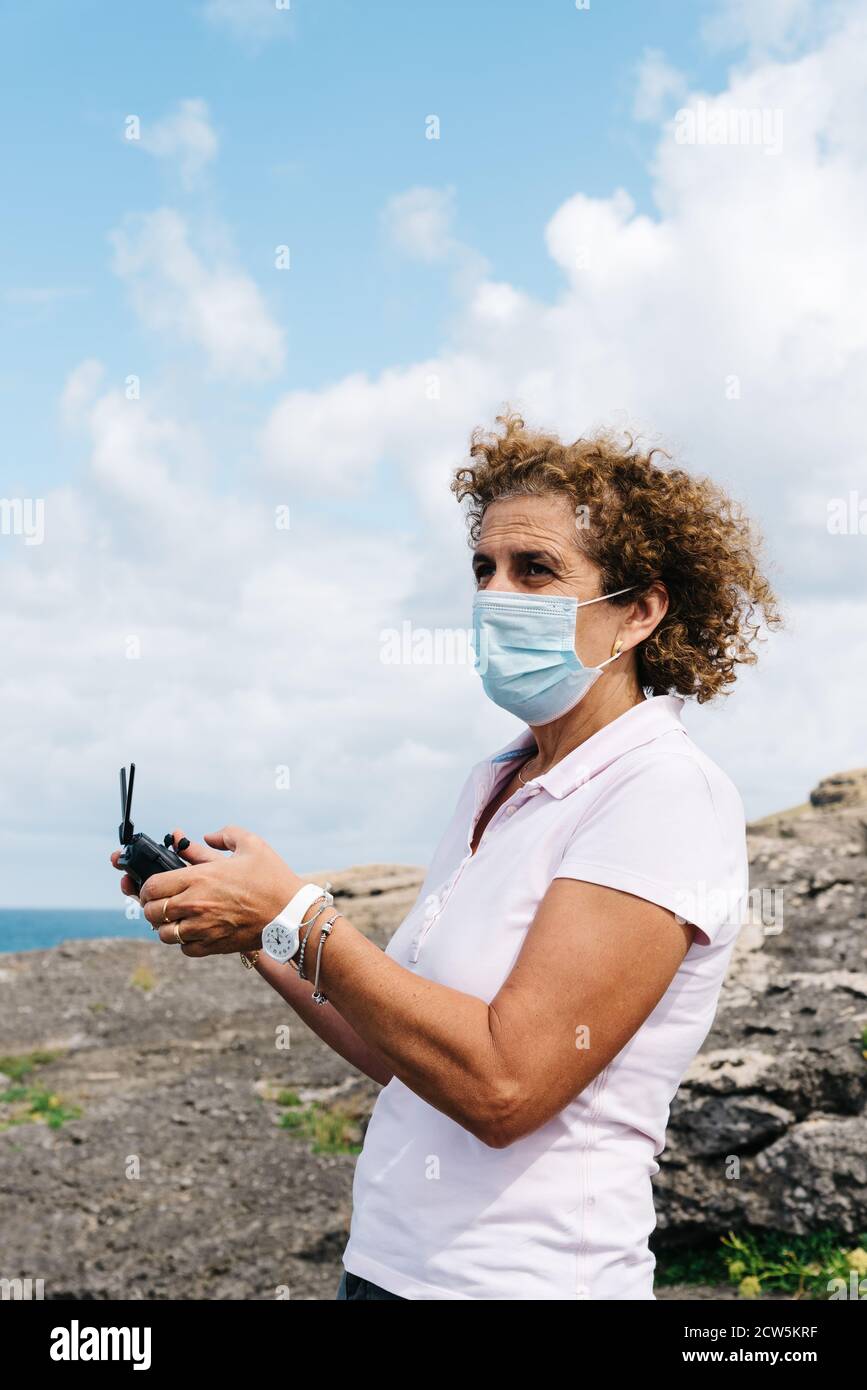 Frau mittleren Alters trägt Gesichtsmaske Navigation einer fliegenden Drohne mit Fernbedienung. Frau in der Natur Drohne fliegen und fotografieren. Stockfoto