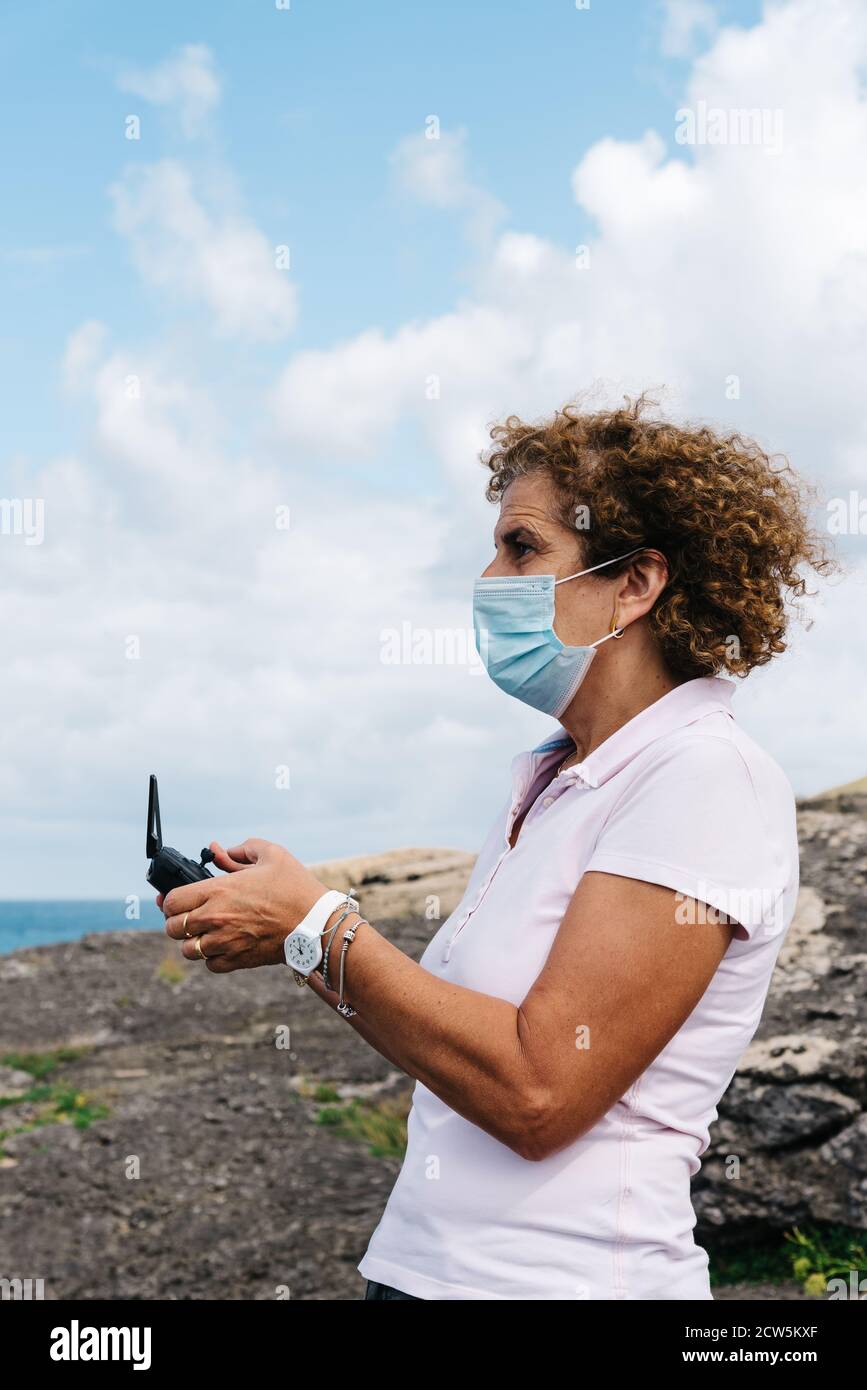Frau mittleren Alters trägt Gesichtsmaske Navigation einer fliegenden Drohne mit Fernbedienung. Frau in der Natur Drohne fliegen und fotografieren. Stockfoto