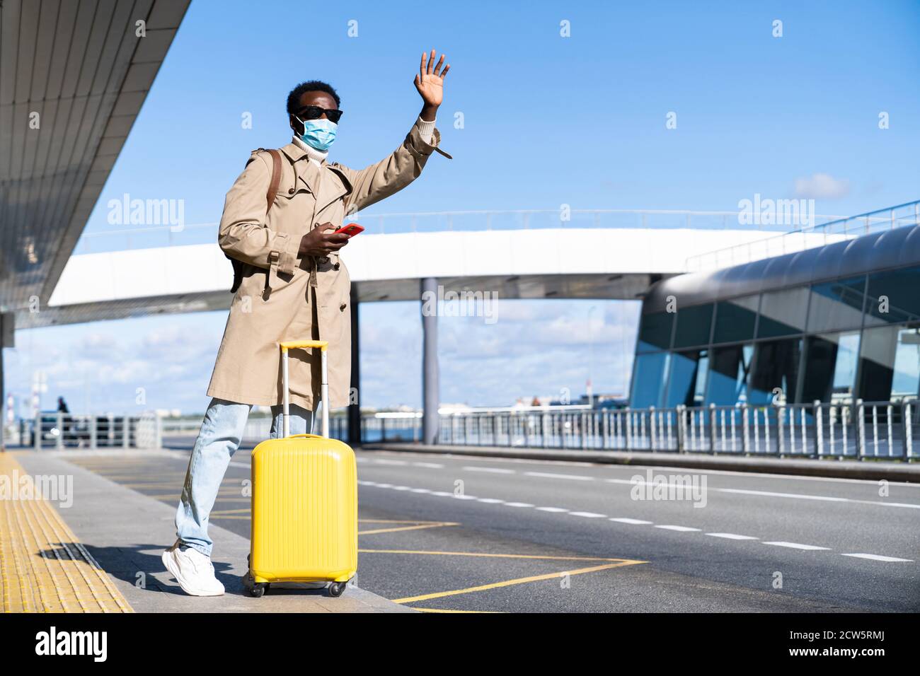 Afro-amerikanische Millennial-Mann mit gelben Koffer steht im Flughafen-Terminal, Telefon, Taxi rufen, Hand heben, Gesicht medizinische Maske zu prote tragen Stockfoto