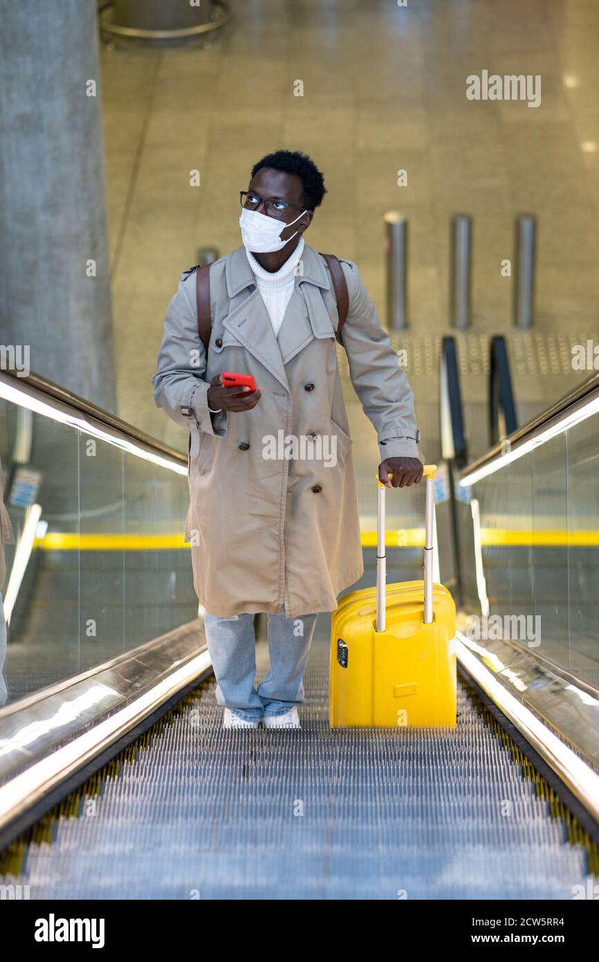 Afro-amerikanischen Reisenden Mann mit gelben Koffer steht auf Rolltreppe in Flughafen-Terminal, tragen Gesicht medizinische Maske, um sich vor Kontakt mit zu schützen Stockfoto