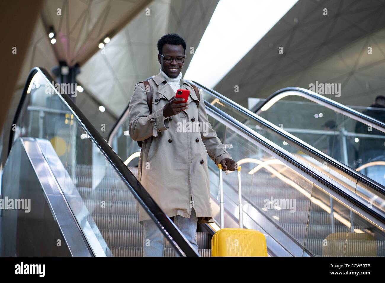 Lächelnder afro-amerikanischer Reisender Mann mit gelben Koffer steht auf Rolltreppe im Flughafenterminal, mit Handy, kommt aus dem Ausland. Stockfoto