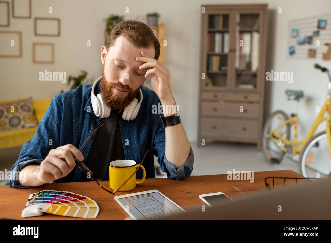 Junge müde männliche freiberufliche Designer berühren Stirn beim Biegen über Becher Stockfoto