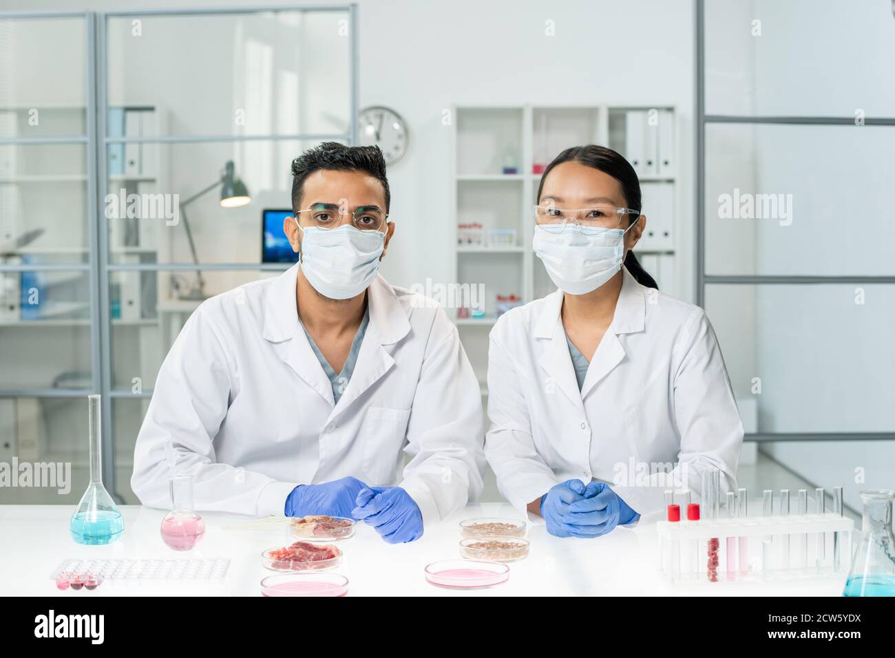 Zwei junge interkulturelle Kliniker in Whitecoats, Schutzmasken und Handschuhen Stockfoto