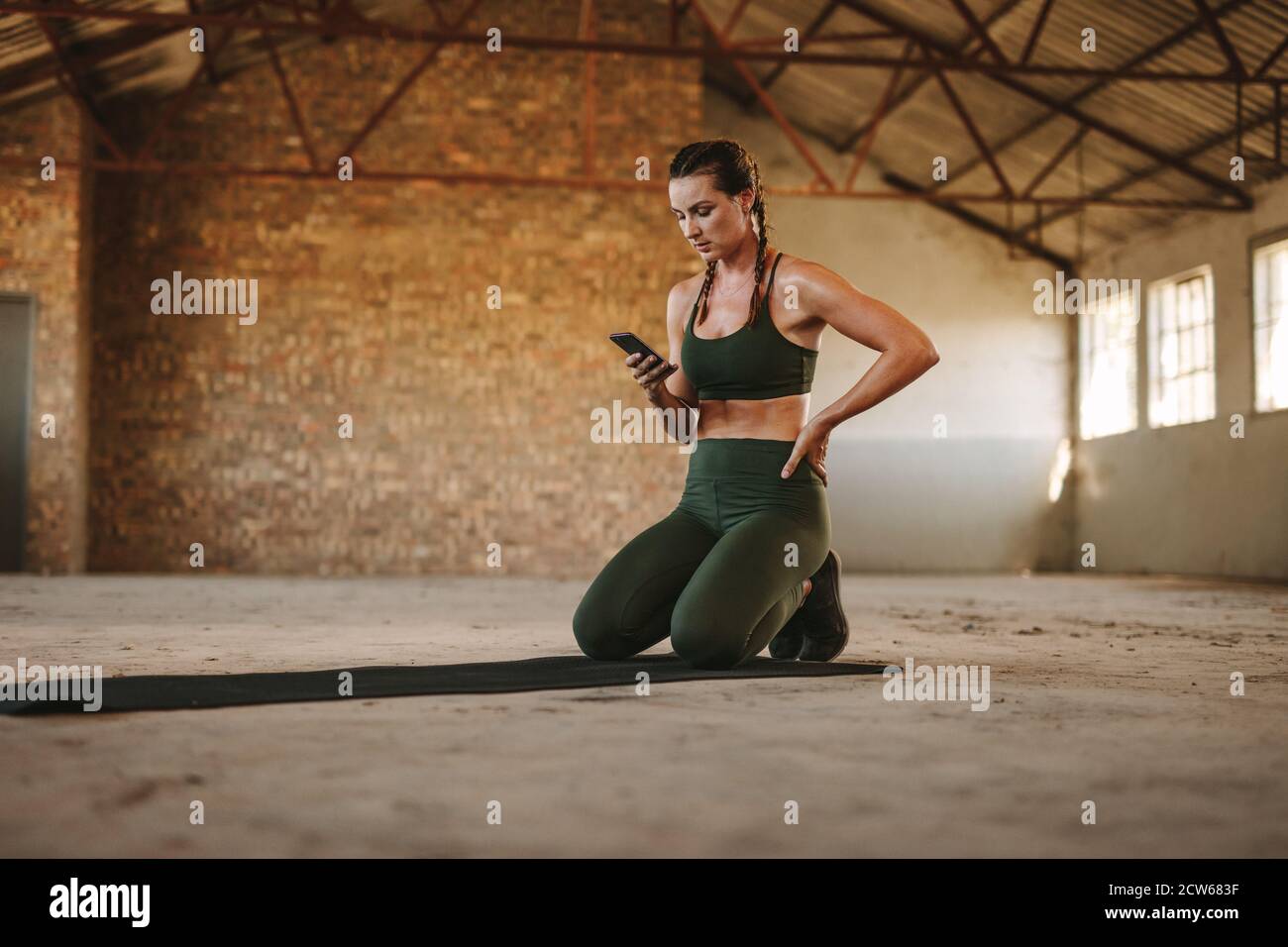 Fitness-Frau, die sich nach dem Training ausruht und das Handy benutzt. Eine Frau sitzt auf einer Trainingsmatte mit einem Handy in einem verlassenen Lagerhaus. Stockfoto